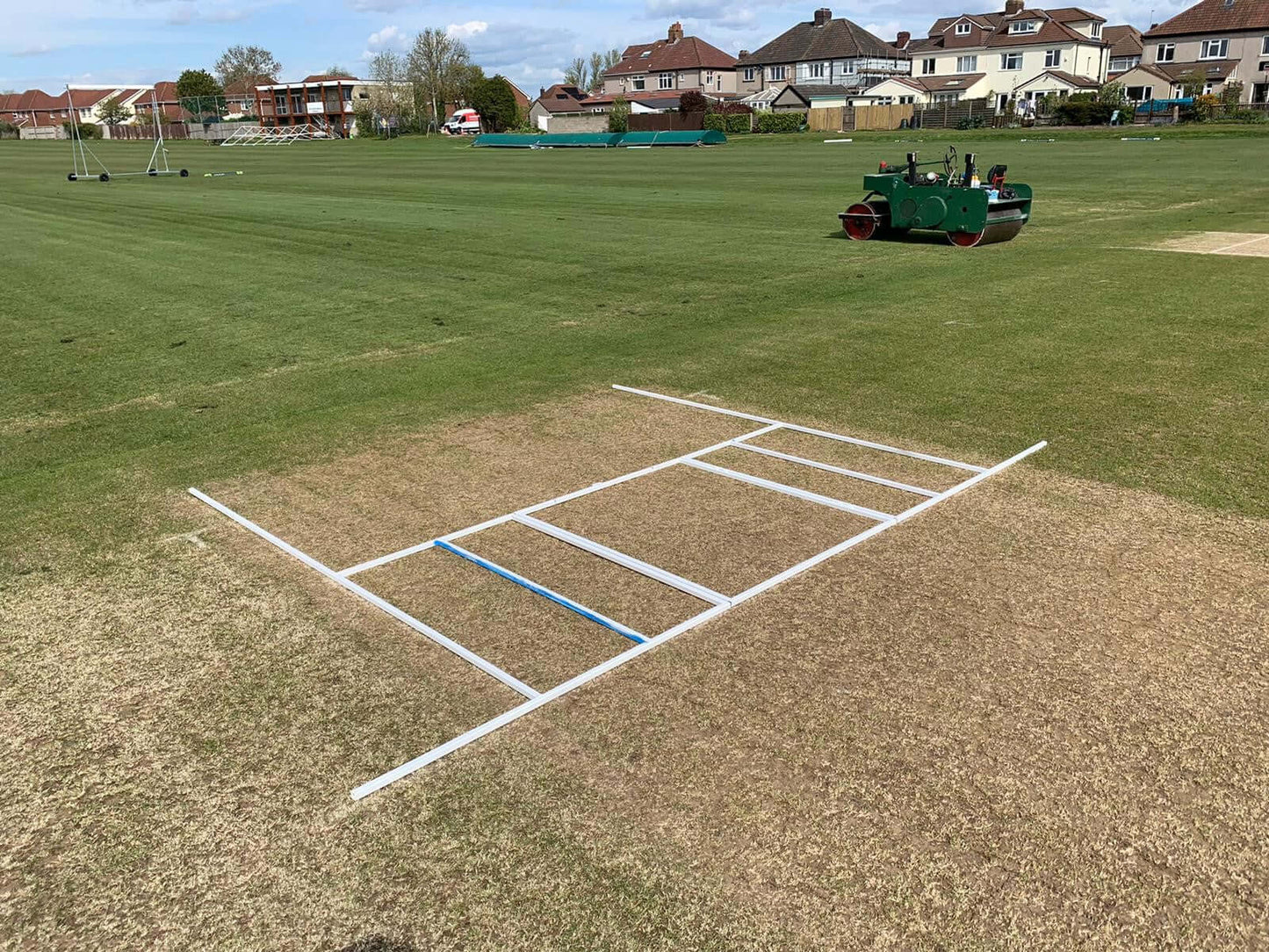 Crease Marking Frame on a cricket pitch with surrounding green grass and homes in the background.