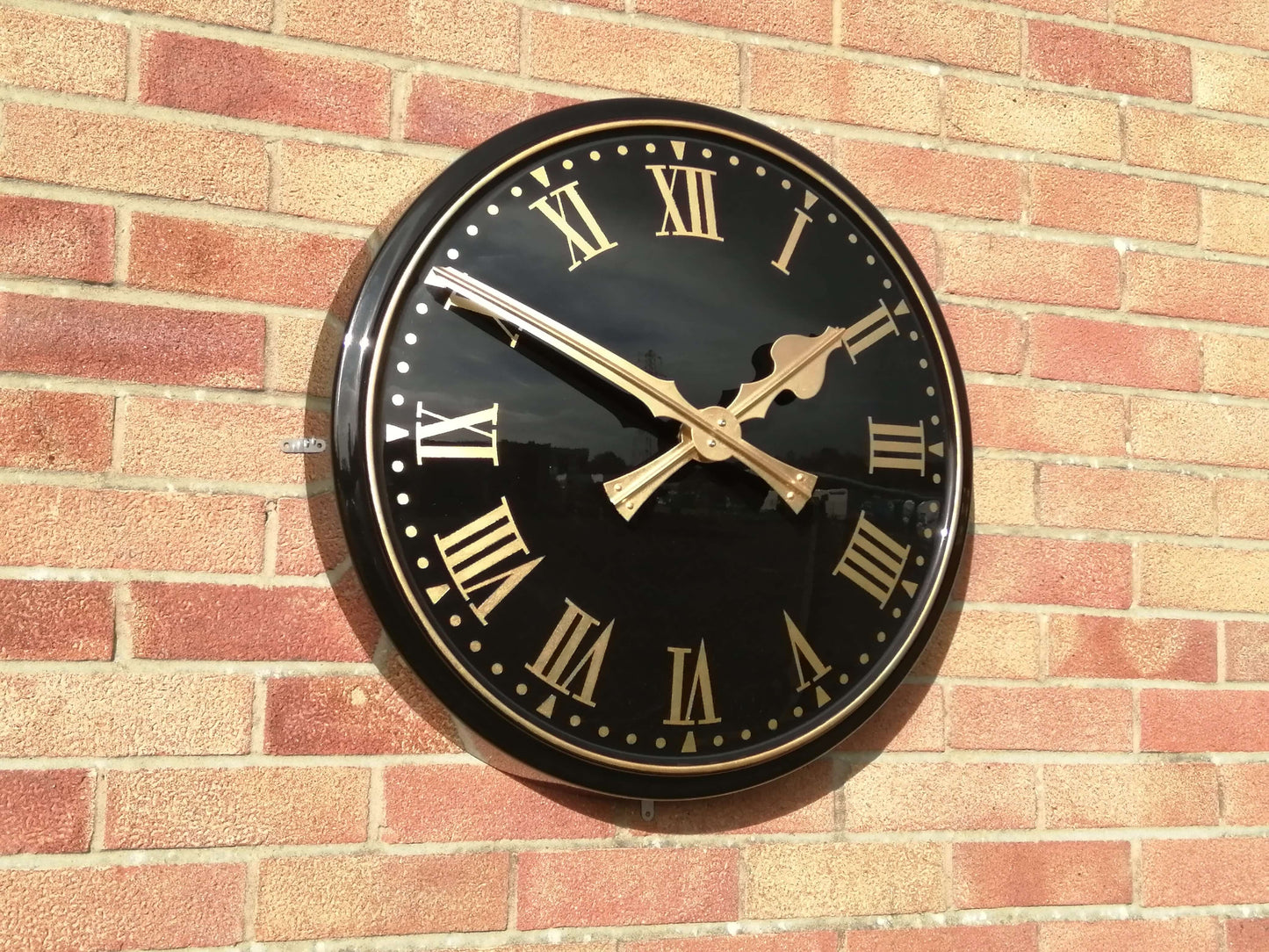 Cricket Pavilion Clocks with black dial and gold hands mounted on a brick wall.